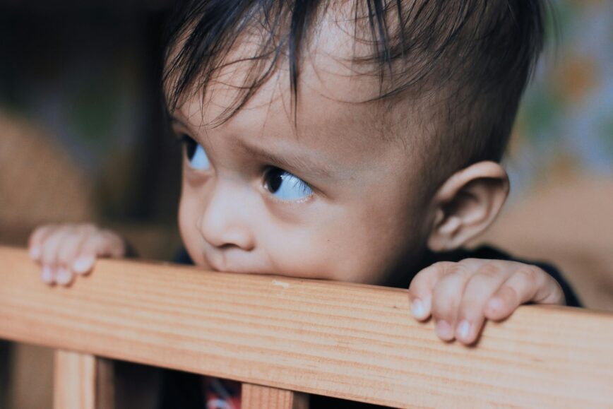 Adorable toddler biting a wooden crib rail, showcasing innocence and curiosity indoors.
