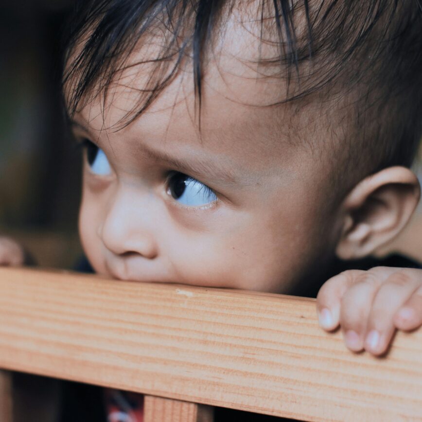 Adorable toddler biting a wooden crib rail, showcasing innocence and curiosity indoors.